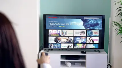 Eine Frau zeigt mit einer Fernbedienung in Richtung eines eingeschalteten Smart-TVs.