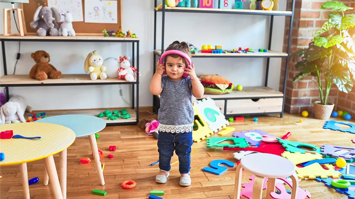 Ein Kind mit rosafarbenen Kopfhörern steht in einem Kinderzimmer. Puzzlematten und Spielzeug liegen auf dem Holzboden.