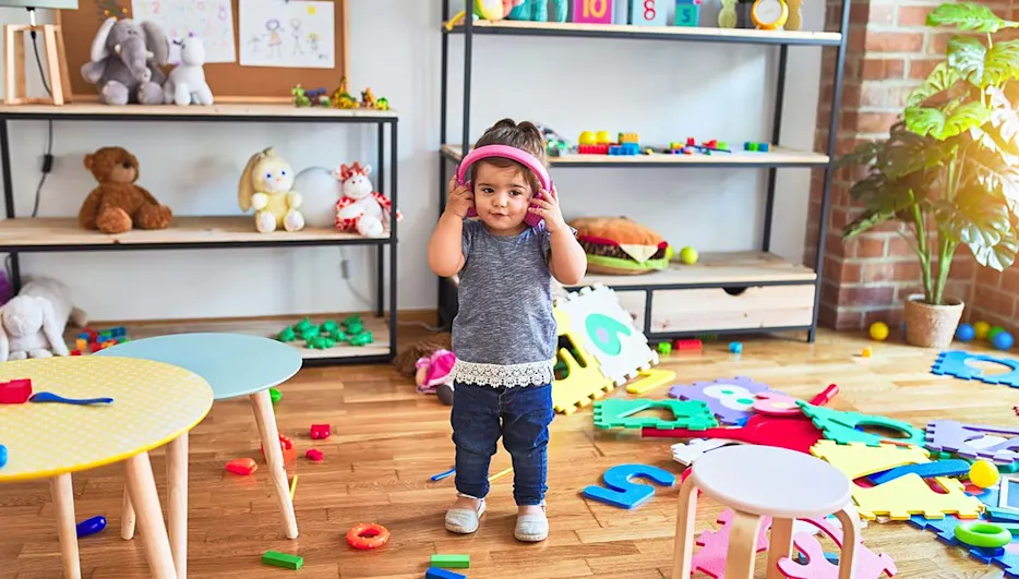 Ein Kind mit rosafarbenen Kopfhörern steht in einem Kinderzimmer. Puzzlematten und Spielzeug liegen auf dem Holzboden.