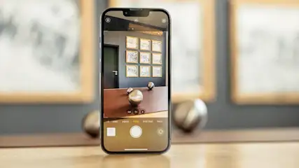 Ein Smartphone zeigt ein Kamerabild mit einer braunen Kommode, silbernen Kugeln und neun gerahmten Bildern an einer grauen Wand.
