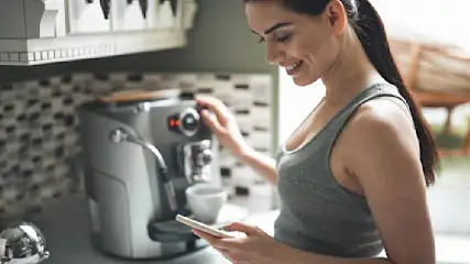 Frau bedient Kaffeemaschine, hält Smartphone. Küche mit gefliester Rückwand, weißen Schränken im Hintergrund.