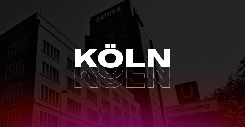 Ein großes, braunes Backsteingebäude mit vielen Fenstern und dem Schriftzug "SATURN" ist zu sehen. Ein Hochhaus befindet sich im Hintergrund.