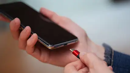 Eine Hand hält ein schwarzes Smartphone, während die andere ein rotes Ladekabel an den Ladeanschluss des Geräts anschließt.