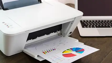 Ein Drucker druckt ein Blatt Papier mit einem farbigen Tortendiagramm. Daneben steht ein Laptop auf einem dunklen Tisch.