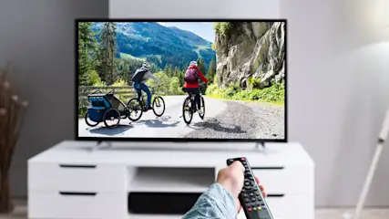 Ein Fernseher zeigt zwei Radfahrer auf einer Bergstraße. Eine Hand hält eine Fernbedienung vor einem weißen Schrank.