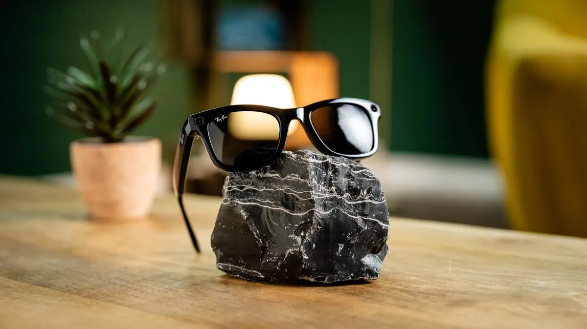 Eine schwarze Brille liegt auf einem dunklen Stein auf einem Holztisch, im Hintergrund sind eine Topfpflanze und ein gelber Sessel.
