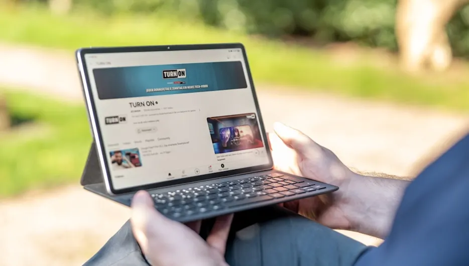 Eine Person hält ein Tablet mit Tastatur im Freien. Auf dem Bildschirm ist eine Webseite zu sehen. Grüner Hintergrund.