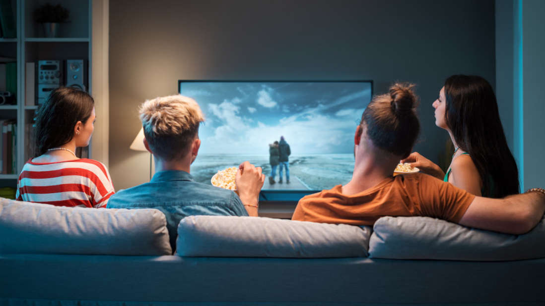 Zwei Männer und zwei Frauen sitzen im Wohnzimmer auf der Couch und schauen gemeinsam einen Film.