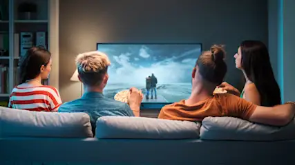 Vier Personen sitzen auf einem Sofa und schauen auf einen Fernseher. Sie essen Popcorn. Im Hintergrund befinden sich ein Bücherregal und eine Wand.