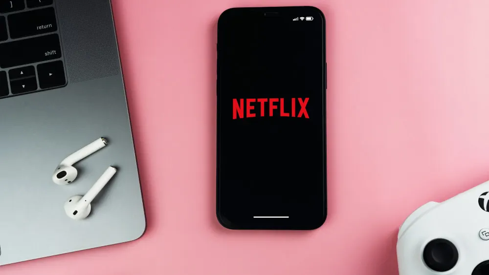 Auf einem rosa Untergrund liegen ein Laptop, Ohrhörer, ein Smartphone mit rotem Netflix-Logo und ein weißer Controller.