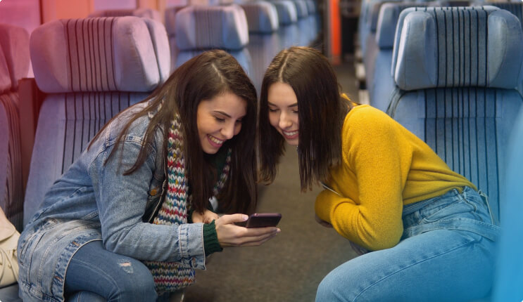 Zwei Frauen sitzen in einem Zug und schauen auf ein Handy. Die Sitze sind blau gestreift.