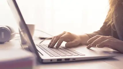 Eine Person tippt auf einem Laptop. Kopfhörer und eine Tasse stehen auf dem Tisch.
