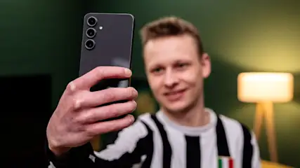 Ein Mann im gestreiften Hemd hält ein schwarzes Smartphone mit drei Kameras vor einem grünen Hintergrund. Eine Lampe ist im Hintergrund.