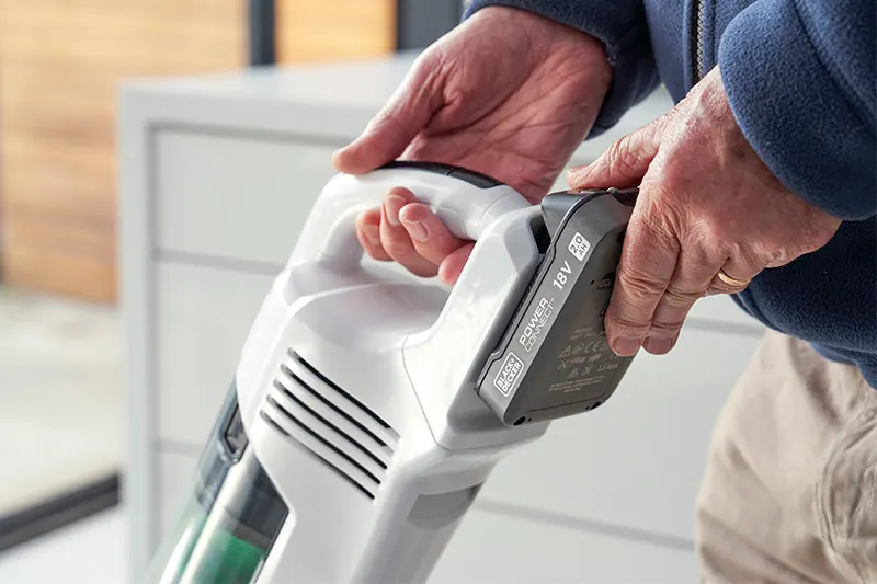 Eine Person hält einen weißen Akku-Staubsauger mit einem grauen 18V-Akku vor einem weißen Schrank.