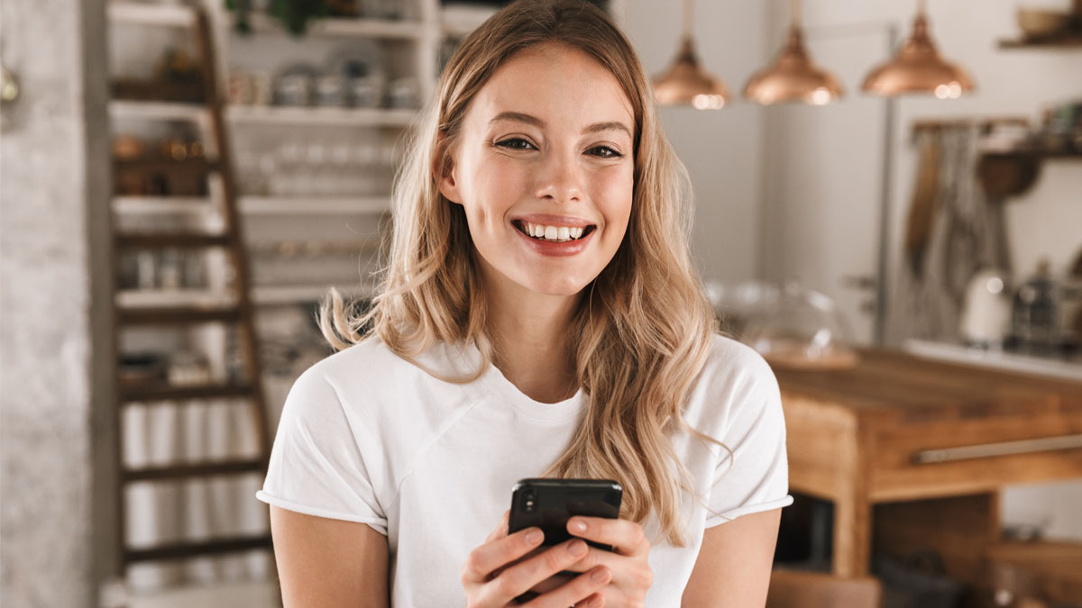 Eine blonde Frau hält ein Smartphone in der Hand und lächelt.