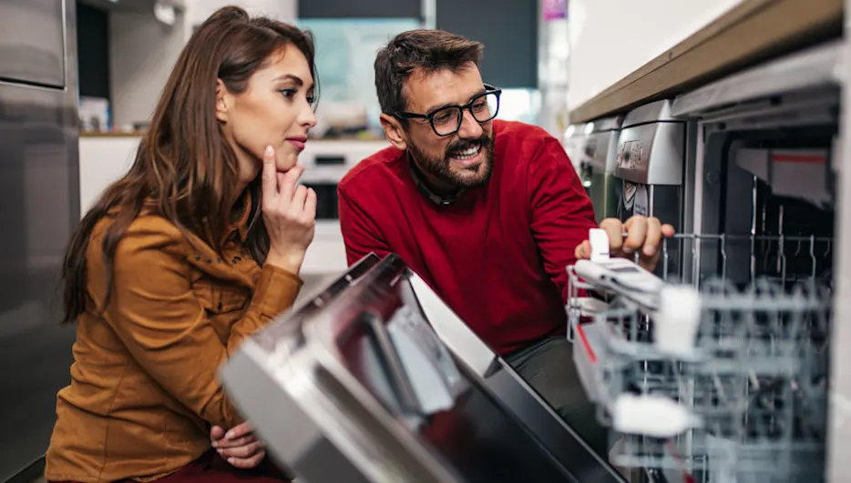 Ein Mann und eine Frau betrachten einen Geschirrspüler in einem Geschäft. Der Mann trägt eine rote Jacke und die Frau eine braune Jacke.