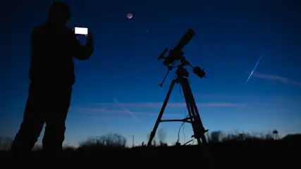 Silhouette einer Person, die ein Telefon hält, und eines Teleskops unter einem blauen Nachthimmel mit Mond und Sternschnuppe.