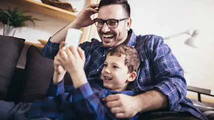 Vater und Sohn lachen auf einer Couch, während sie ein Smartphone betrachten. Beide tragen karierte Hemden.
