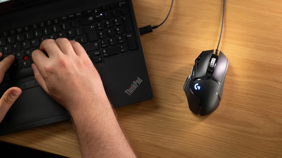 Die Logitech G502 Hero liegt rechts neben einem schwarzen ThinkPad-Laptop, auf dem Hände tippen.