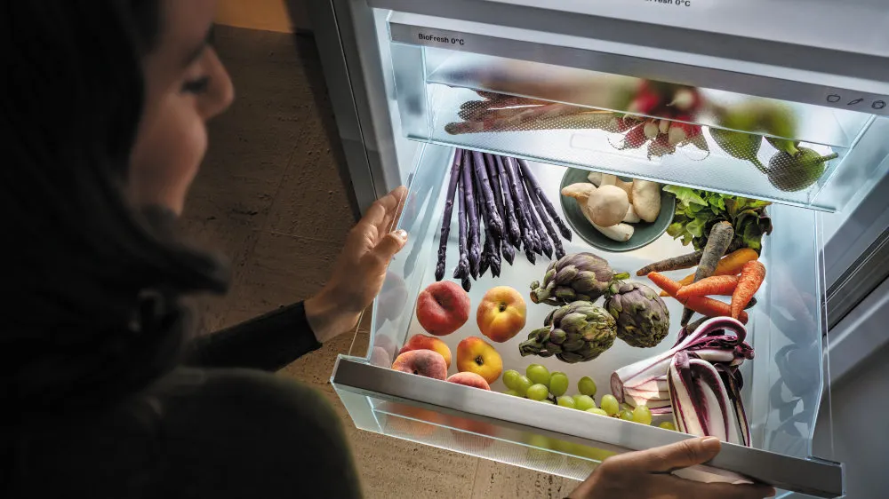 Eine Frau öffnet einen Kühlschrank, der mit Obst und Gemüse wie Spargel, Pfirsichen, Trauben und Karotten gefüllt ist.