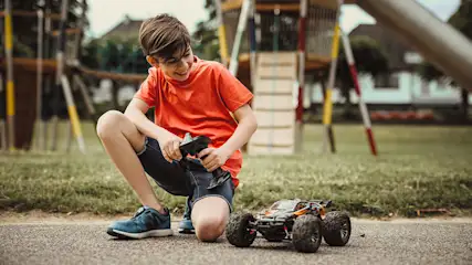 Ein Junge mit orangefarbenem T-Shirt hockt mit einer Fernbedienung neben einem Spielzeugauto im Park. Im Hintergrund ist ein Spielplatz.