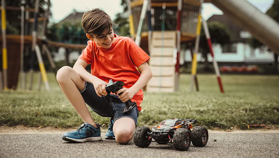 Ein Junge mit orangefarbenem T-Shirt hockt mit einer Fernbedienung neben einem Spielzeugauto im Park. Im Hintergrund ist ein Spielplatz.