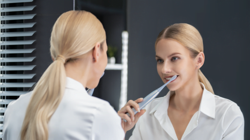 Eine Frau in weißem Hemd putzt sich vor einem Spiegel mit einer blauen elektrischen Zahnbürste die Zähne. Jalousien im Hintergrund.