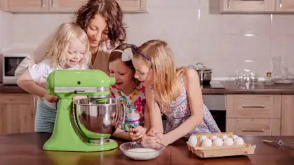 In der Küche schauen eine Frau und drei Mädchen auf einen grünen Mixer, Eier und eine Schüssel mit Zucker auf einem Holztisch.