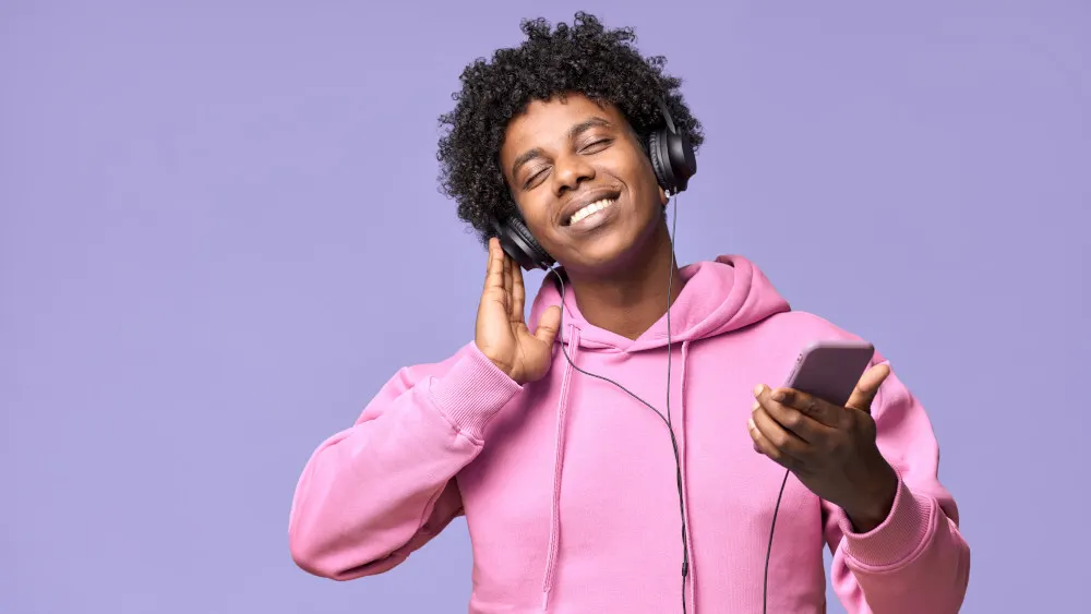 Mann mit Kopfhörern und rosa Kapuzenpullover hält ein Telefon vor einem lila Hintergrund.