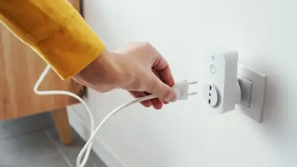 Eine Hand mit gelbem Ärmel steckt einen weißen Stecker in eine weiße Steckdose an einer weißen Wand. Ein Holzmöbelstück ist im Hintergrund.