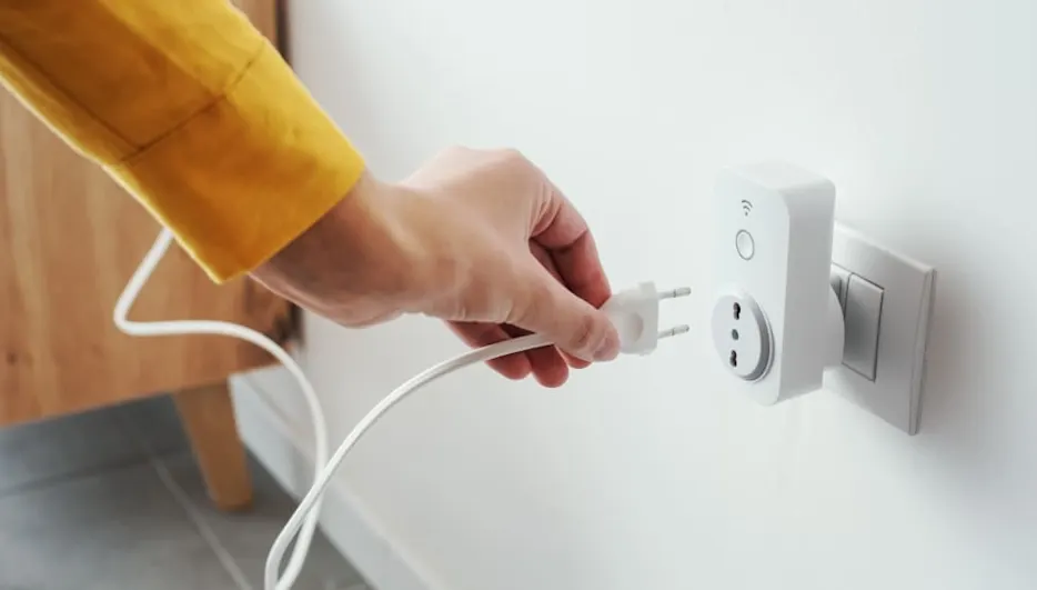 Eine Hand mit gelbem Ärmel steckt einen weißen Stecker in eine weiße Steckdose an einer weißen Wand. Ein Holzmöbelstück ist im Hintergrund.