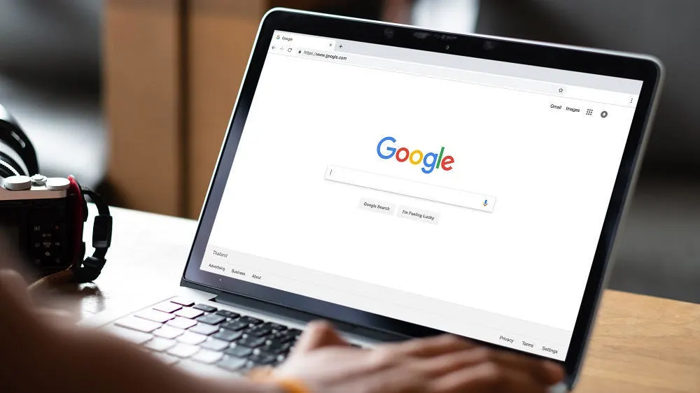 Ein Laptop zeigt die Google-Startseite mit Suchleiste. Eine Kamera und Kopfhörer stehen daneben auf einem Holztisch.