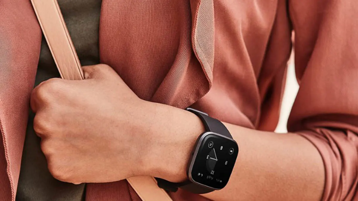 Eine Frau im Blazer trägt eine cremefarbene Umhängetasche und hält mir ihrer Hand den Riemen fest. Am Handgelenk trägt sie eine Fitbit Versa 3 in schwarz.