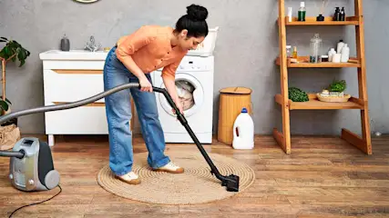 Frau saugt einen runden Teppich auf einem Holzboden. Im Hintergrund steht eine Waschmaschine.