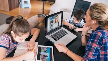 Auf dem Tisch sitzen Frau, Mädchen, Junge mit Laptop, Tablet und Notizbuch. Das Mädchen schreibt, der Junge spielt und die Frau hat eine Videoanruf.