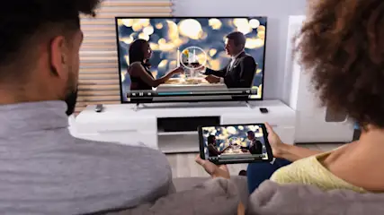 Ein Paar sitzt vor einem Fernseher und einem Tablet, auf denen ein Mann und eine Frau zu sehen sind, die anstossen.