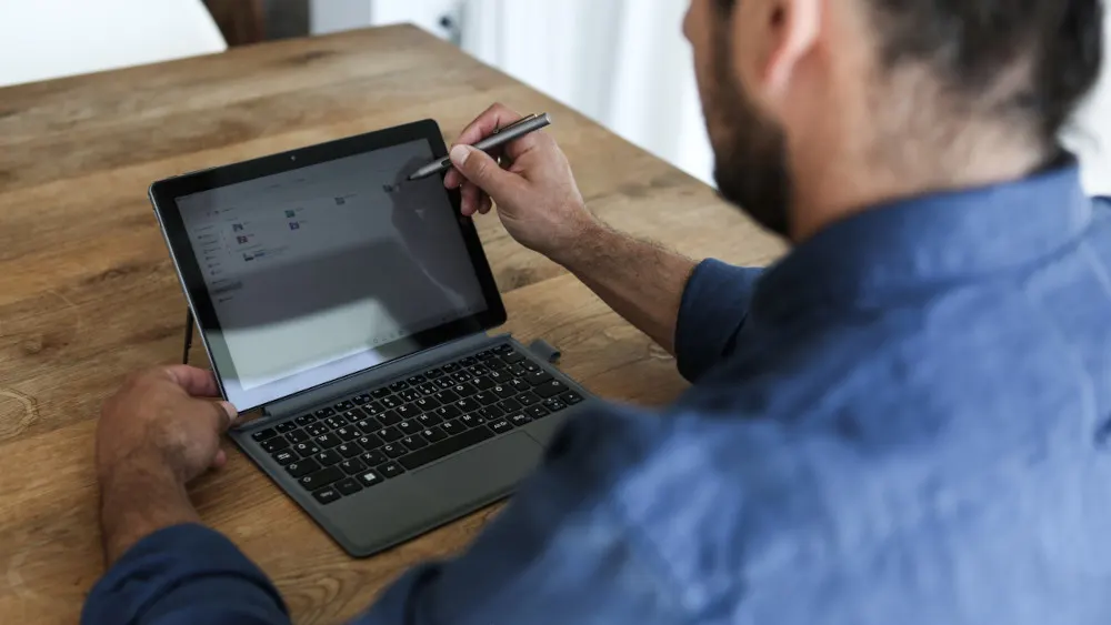 Ein Mann sitzt an einem Holztisch und benutzt einen Stift auf einem Tablet-Computer mit Tastatur.