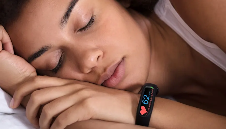 Eine schlafende Person mit einem schwarzen Fitness-Tracker am Handgelenk, der eine Herzfrequenz von 62 anzeigt.