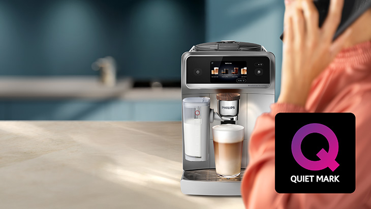 Hintergrund: Kaffeemaschine. Vordergrund: Eine Frau telefoniert mit ihrem Handy. Ein Logo mit der Aufschrift "Quiet Mark" ist in der Ecke abgebildet.