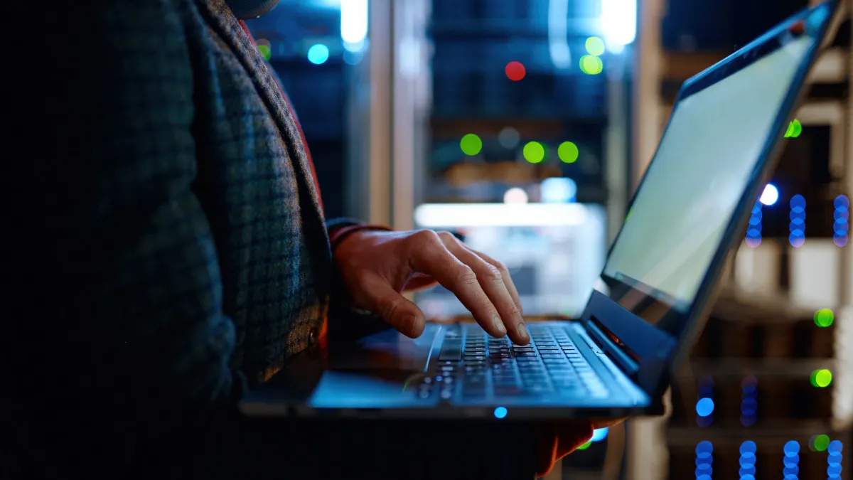 Eine Person tippt auf einem Laptop vor einem Serverschrank mit blinkenden Lichtern. Der Laptop ist geöffnet und beleuchtet.