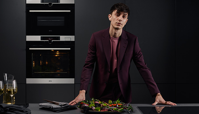 Ein Mann in einem burgunderfarbenen Blazer steht in einer Küche mit schwarzen Schränken neben einem Doppelbackofen und einem Teller Salat.