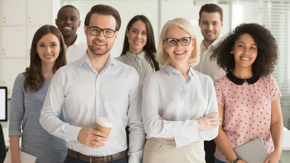 Eine Gruppe von acht lächelnden Personen steht in einem Büro und trägt Hemden und Blusen. Ein Mann hält einen Kaffeebecher.