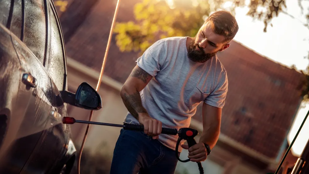Ein Mann in Jeans und T-Shirt wäscht mit einem Hochdruckreiniger ein Auto im Freien. Im Hintergrund sind Bäume und ein Haus.