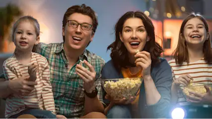 Eine Familie, bestehend aus Eltern und zwei Töchtern, sitzt zusammen und isst Popcorn vor einem hellen Bildschirm.