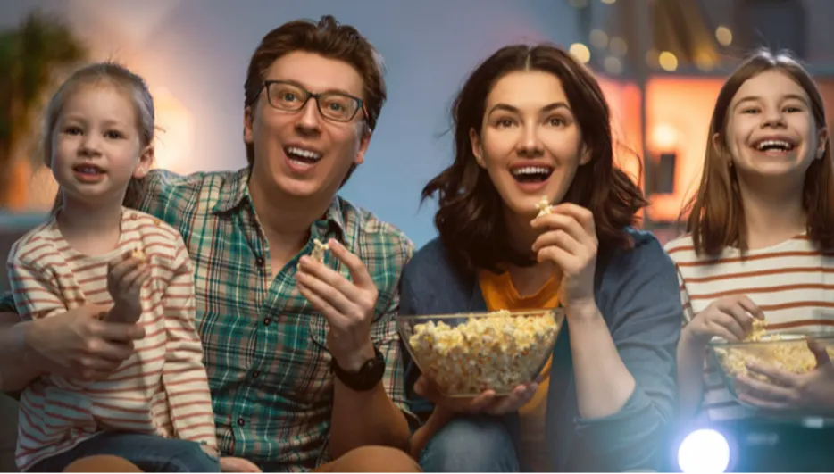 Eine Familie, bestehend aus Eltern und zwei Töchtern, sitzt zusammen und isst Popcorn vor einem hellen Bildschirm.