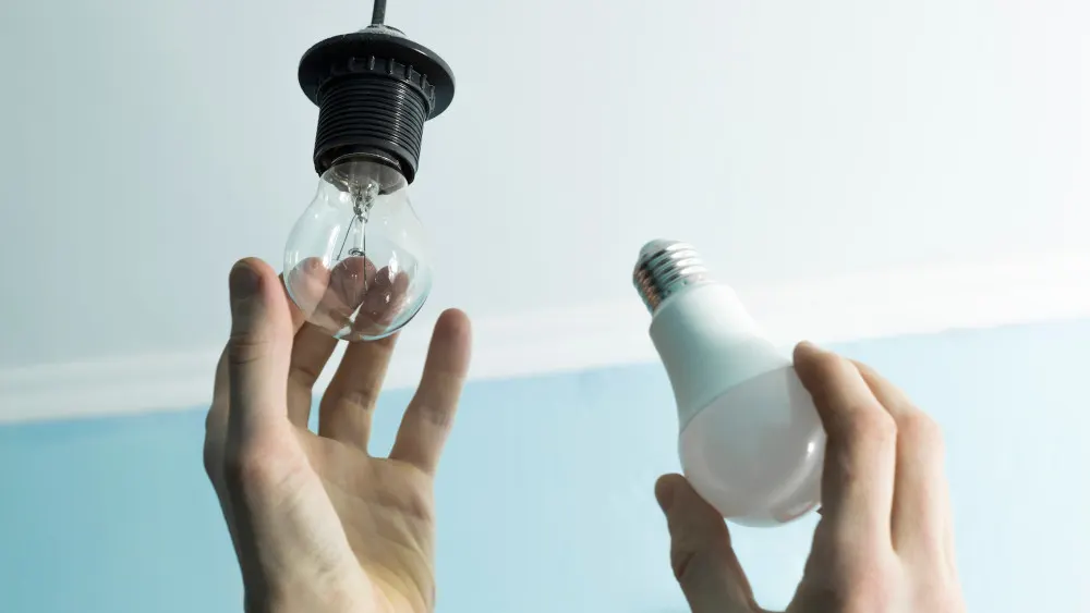 Eine Hand hält eine klare Glühbirne unter einer schwarzen Fassung, die andere eine weiße LED-Glühbirne; hellblauer Hintergrund.