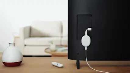 Auf einem Tisch liegen eine Fernbedienung, eine Vase und ein an einen schwarzen Fernseher angeschlossener, weißer Streaming-Stick.