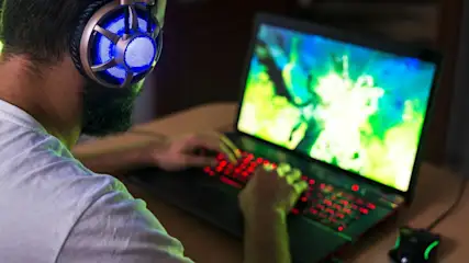 Person mit Kopfhörern tippt auf einem Laptop mit roter Tastatur und Maus auf einem Tisch. Der Bildschirm ist blau und grün.