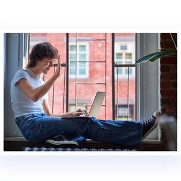 Eine Person sitzt am Fenster mit einem Laptop und einer Pflanze. Die Person trägt ein helles T-Shirt und eine blaue Jeans.
