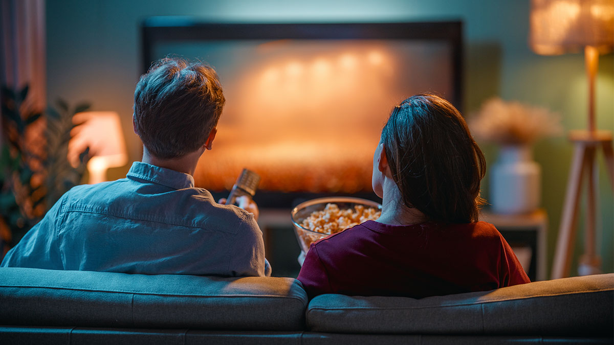 Zwei Personen sitzen auf dem Sofa vor dem Fernseher. Eine Person hält eine Fernbedienung, die zweite eine Schüssel Popcorn.